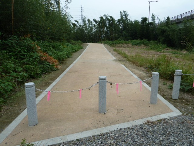灰色のポールとチェーンで封鎖された舗装路が草木に囲まれ奥へまっすぐ延びる様子を写した写真