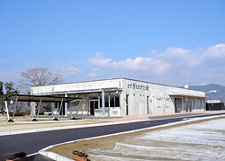 青空の下、平屋のコンクリート建物が山並みを背に日差しを受けつつ広い舗装路と芝生に囲まれて佇むさが水ものがたり館の外観写真