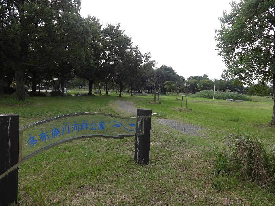 緑の芝生と並木が広がる静かな公園入口で「多布施川河畔公園」の木製看板が小径脇に立つ写真