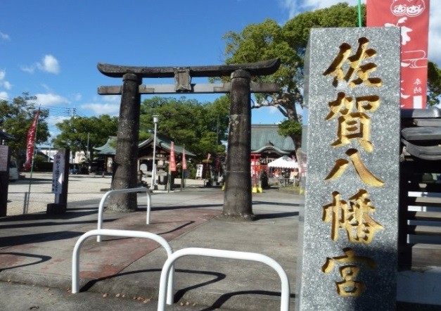 青空の下、「佐賀八幡宮」と刻まれた石碑の横に古い鳥居が立ち、境内の社殿や旗が奥に続く情景の写真
