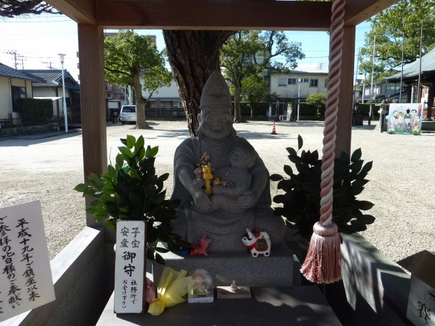 木造の覆いの下で子どもを抱く子育て恵比須像の前に葉飾りや小さなお供え物が並び静かに祀られている写真