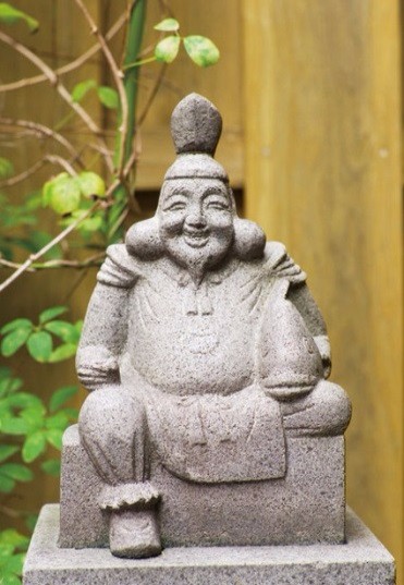 木の塀の前に置かれた衣装をまとった恵比須さんの石像が台座に静かに腰掛けている写真