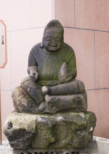 石造の恵比須さんの座像が岩状の台座に穏やかに腰掛け、淡い光に照らされて佇む姿の写真