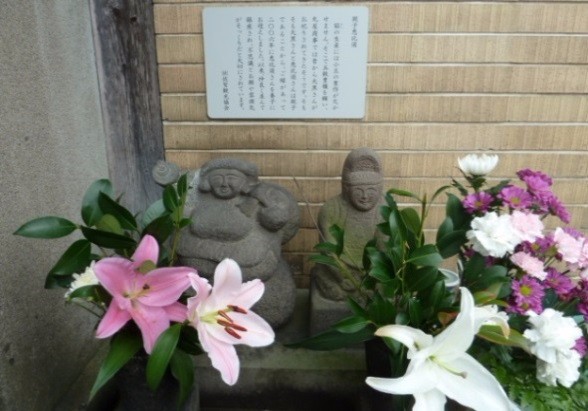 石像二体が色鮮やかな百合や菊など多様な供花に囲まれ、背後の説明板の下で静かに祀られている円満恵比須の写真
