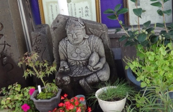 鉢植えの草花や茂る緑に囲まれて屋外の一角に静かに佇み、背後の額装掲示物と木製壁が調和する恵比須像の写真