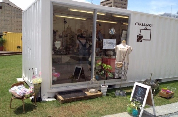 白いコンテナ店舗の前にマネキンや鉢植え、雑貨や黒板風看板が整えられ芝生に囲まれて佇む様子の写真