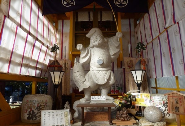 釣竿を掲げ鯛を抱えたとんさん恵比須が灯籠や花、案内札と共に飾られ、柔らかな光に包まれた社殿内で厳かに佇む写真