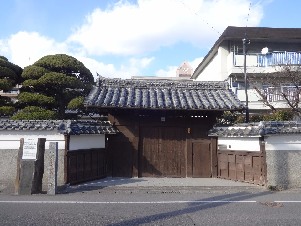 木造瓦屋根の伝統的な門が整えられた庭木と白壁に囲まれ武家屋敷の門を正面から撮影した写真