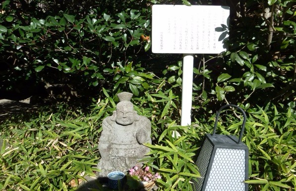 木々が茂る庭の一角に小さなあけぼの宝珠恵比須像と案内板が立ち、手前に供え物の器や灯籠が並び光を受けて静かに佇む様子を丁寧に写した写真