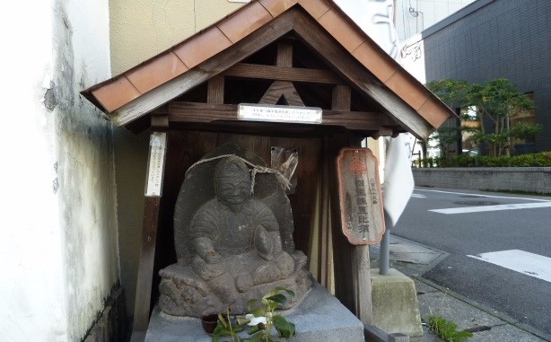 道路脇の小さな木製祠に安置された石像が古びた札とともに佇み、背景に街並みが見える写真