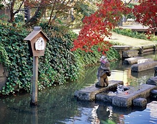 紅葉が水辺を彩る中、水面上の石に立つ小像と木製案内板が並び穏やかな情景をつくる写真