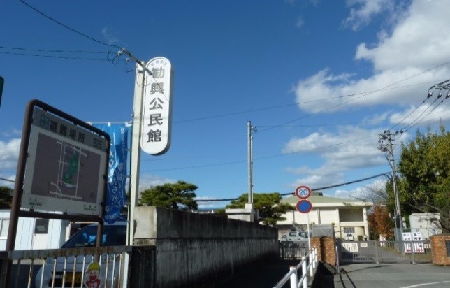 青空の下、「公民館」の縦看板と案内板が道路脇に立ち、奥に建物や樹木が並ぶ街角の様子の写真