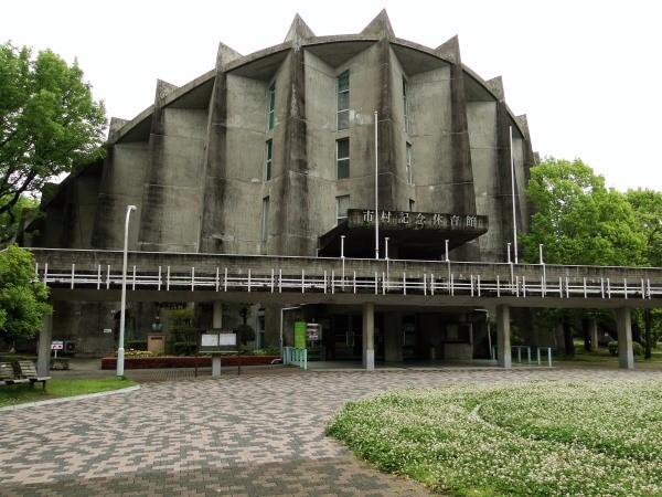 コンクリート造の独特な多角形外観を持つ大きな建物が並木に囲まれた舗道の先に堂々と建つ市村記念体育館の外観写真