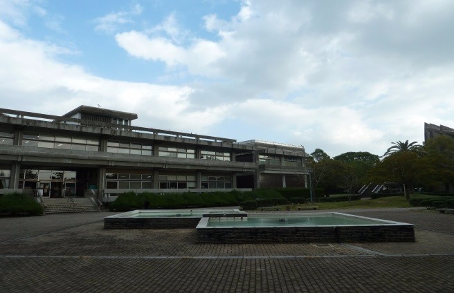 広い中庭に浅い水盤が並び、その奥に直線的なコンクリート造の建物が立つ佐賀県立図書館の外観写真