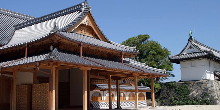青空の下、瓦屋根と木組みが端正に連なる城郭建築が広がり、石垣と豊かな緑が景観に重厚さを添え、落ち着いた雰囲気を醸し出す佐賀城本丸歴史館の外観写真