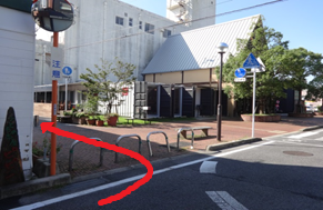 街角で建物横の歩道へ赤い矢印が左方向への進行を示し、広場と樹木が並ぶ景観を写した写真