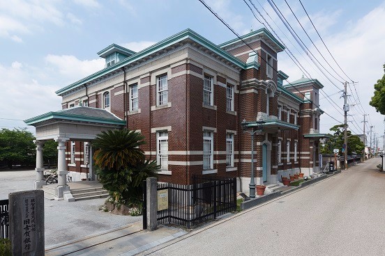 赤茶色のレンガと白い装飾が特徴的な旧古賀銀行が道路沿いに堂々と建ち周囲の緑と調和する写真