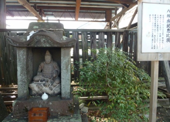 覆い屋根の下で石造の像が祠に安置され、横の説明板と茂る植栽が写っている写真