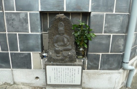 黒いタイル壁の窪みに石像が据えられ、その横に榊の枝葉と小さな供物と説明板が揃えて置かれた松川屋恵比須の写真