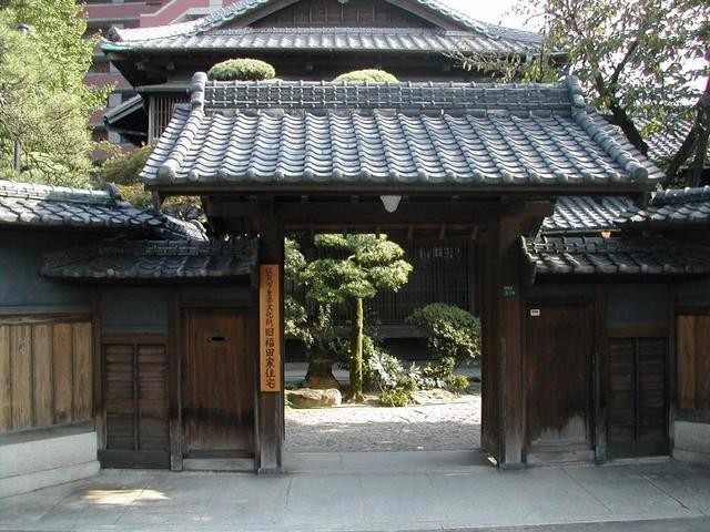 瓦屋根と木の門が落ち着いた佇まいで庭木の緑を囲むように構えた旧福田家の入口を写した写真