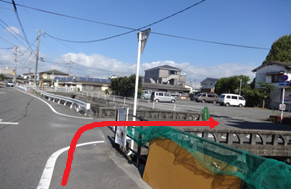 道路沿いの歩道脇で赤い矢印が右折して進む方向を示し、住宅街と駐車場が広がる様子を写した写真
