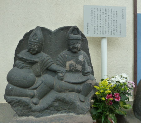 二体のなかよし恵比須の石像が並んで座り横に説明板と色とりどりの供花が添えられている写真
