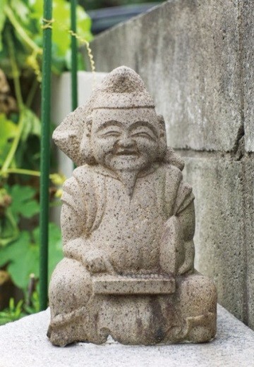 コンクリート壁際に置かれた小さなそろばん恵比須が静かに佇み、背後の緑が柔らかく彩る写真