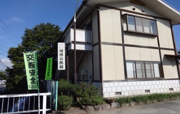 青空の下に立ち、前方には緑の旗や植栽が並ぶ淡い色の外壁を持つ公民館の建物の写真