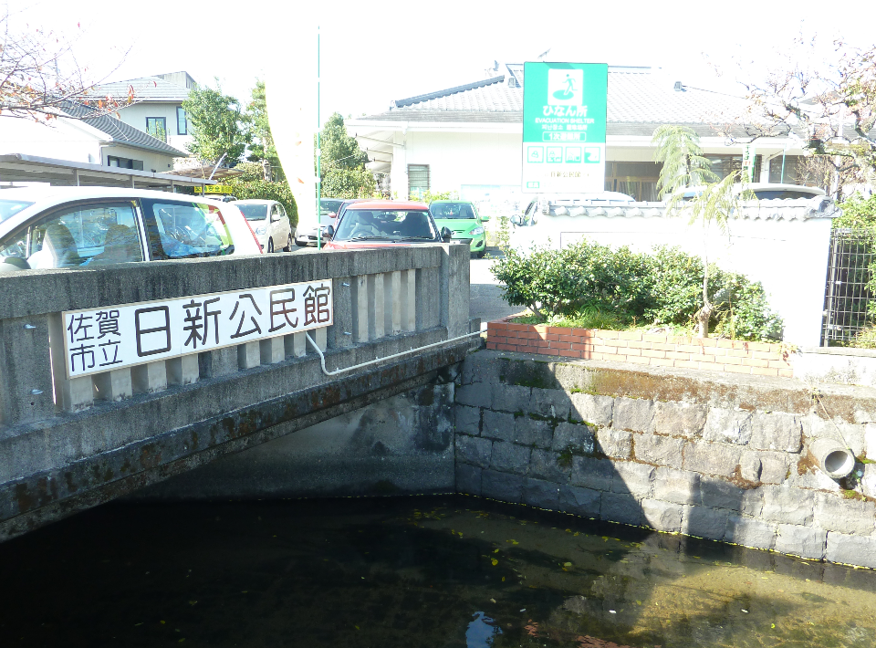 「佐賀市立日新公民館」と書かれた看板が設置されたコンクリート造りの橋のたもとで、石積みの護岸が続く水路を眼下に眺めながら、背後に広がる避難所の看板や駐車場を含めた地域の公共施設周辺の写真
