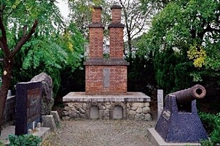 木々に囲まれた屋外の広場に、赤茶色の煉瓦を積み上げた二本の煙突のような特徴的な形状を持つ築地反射炉跡がそびえ立ち、その手前には歴史を感じさせる鉄製の大砲が展示されている、静かな史跡の風景を捉えた写真