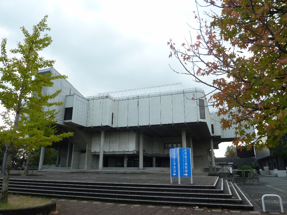 曇り空の下、木々の緑や紅葉に囲まれた近代的な白い外壁のコンクリート建築が建っており、手前の広場には青い案内看板が設置されている佐賀県立博物館の外観を捉えた写真