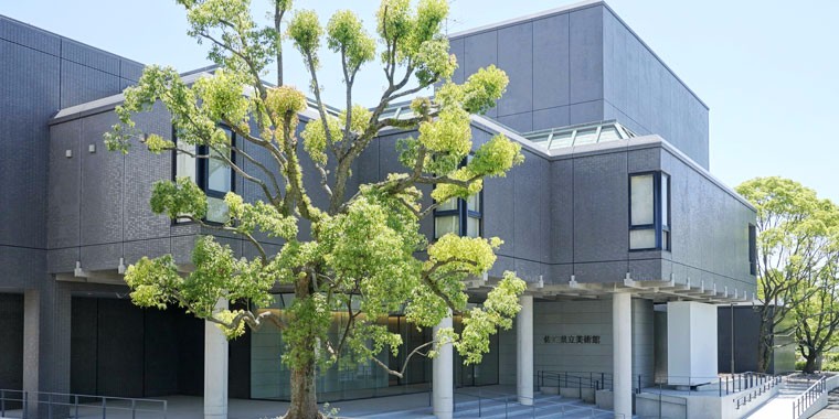 晴天の下で、グレーのタイルの外壁が特徴的な近代的な建物の入り口付近に青々と茂った大きな木が立ち、壁面に「佐賀県立美術館」の文字が刻まれたエントランス周辺の様子を斜め前から捉えた写真