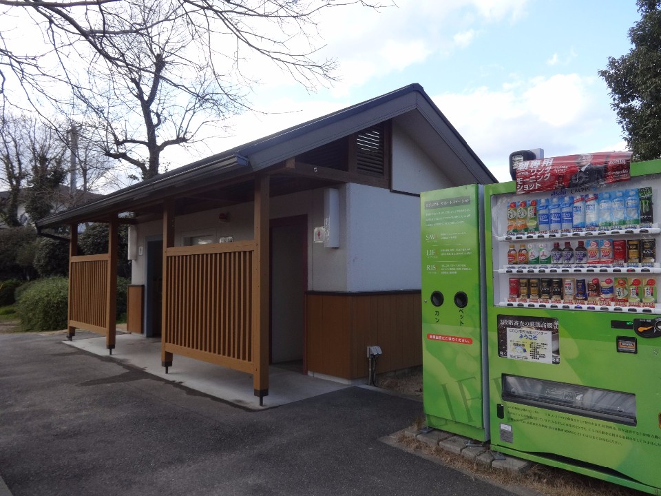 冬の澄んだ空気を感じさせる青空の下、木製の格子の仕切りが特徴的な落ち着いたデザインの公衆トイレが建っており、その手前には緑色の飲料自動販売機と空き缶・ペットボトルの回収ボックスが並んで設置されている公園内の様子を捉えた写真