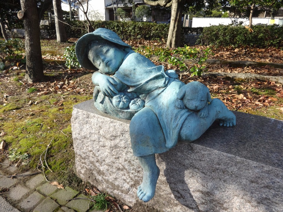 冬の柔らかな日差しが差し込む静かな公園で、帽子をかぶり、果物を入れた袋を枕にして心地よさそうに眠る小さな子供と、その膝の上で丸くなって一緒に眠る子犬の姿を愛らしく表現したブロンズ像を捉えた写真