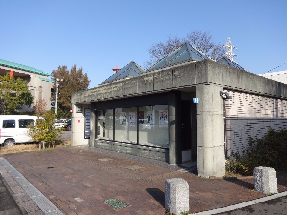 抜けるような青空の下、コンクリート造りの平屋建ての建物の屋根に、ピラミッドのような形をした3つのガラス窓が特徴的に並び、壁面の大きなガラス窓にはポスターが掲示されているトイレの写真