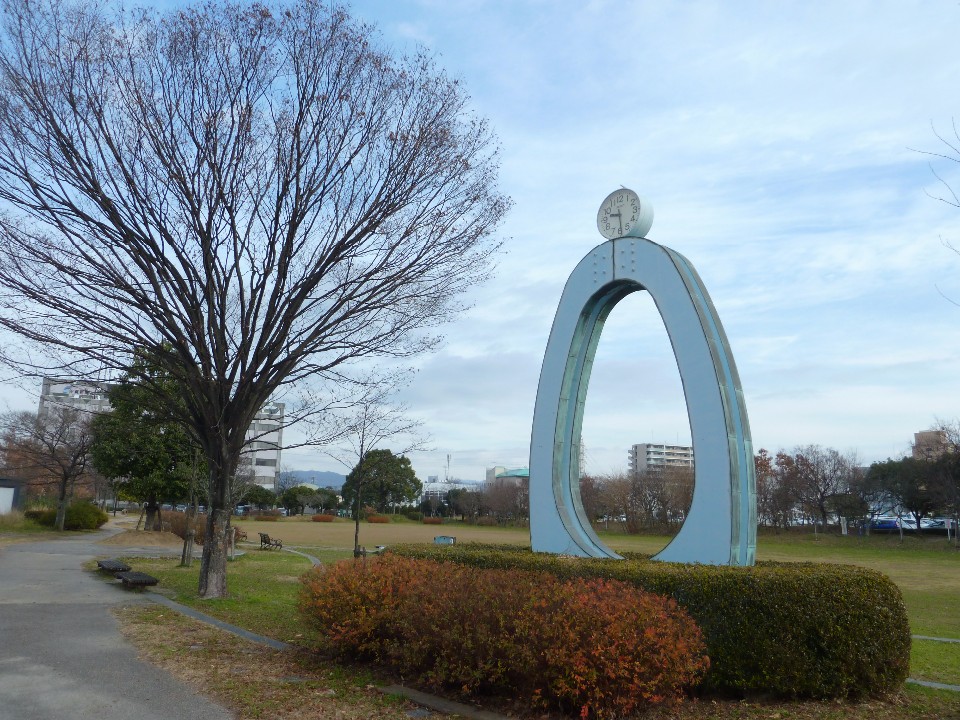 冬の薄曇りの空の下、落葉した大きな樹木が立つ広々とした公園の一角に、水色をした楕円形の巨大なモニュメントが鎮座し、その頂点には円形の時計が設置されている写真