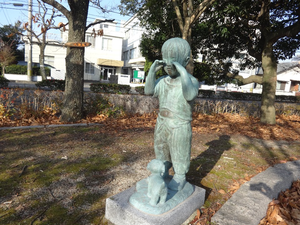 冬の柔らかな日差しが差し込む公園の広場で、木々に囲まれた一角に設置された、目をこすりながら泣いている小さな子どもと足元に寄り添う子犬の様子を表現したブロンズ像を、正面から捉えた写真