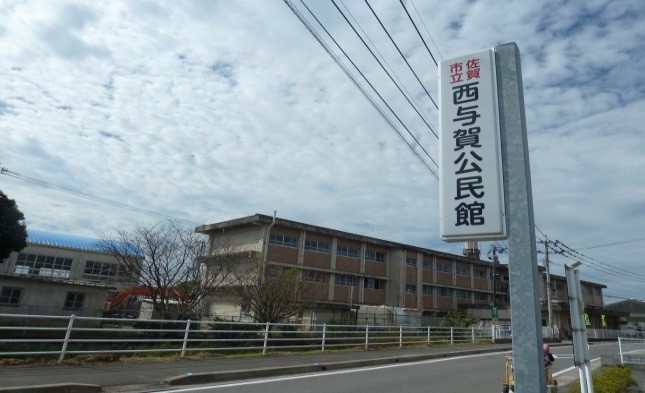 薄曇りの空の下、道路沿いに設置された「佐賀市立西与賀公民館」と書かれた縦長の白い看板の写真