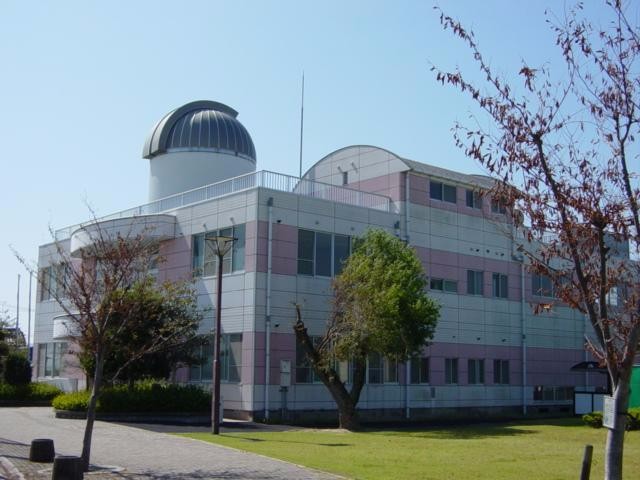 晴れ渡る青空の下、白と薄いピンク色の外壁が特徴的な三階建ての建物の屋根に、天体観測用と思われる銀色のドーム型施設が設置された「佐賀市立星空学習館」を、手前の緑豊かな芝生広場とともに捉えた写真