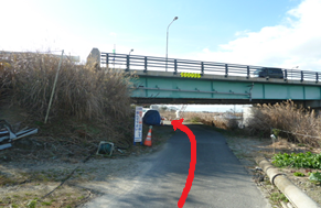 冬の澄み渡る青空の下、枯れ草が広がる河川敷の細い未舗装路を進み、前方の大きな緑色の鉄橋の下をくぐり抜けてさらに先へと続くルートを、路面に合成された大きな赤い矢印で分かりやすく示した風景写真