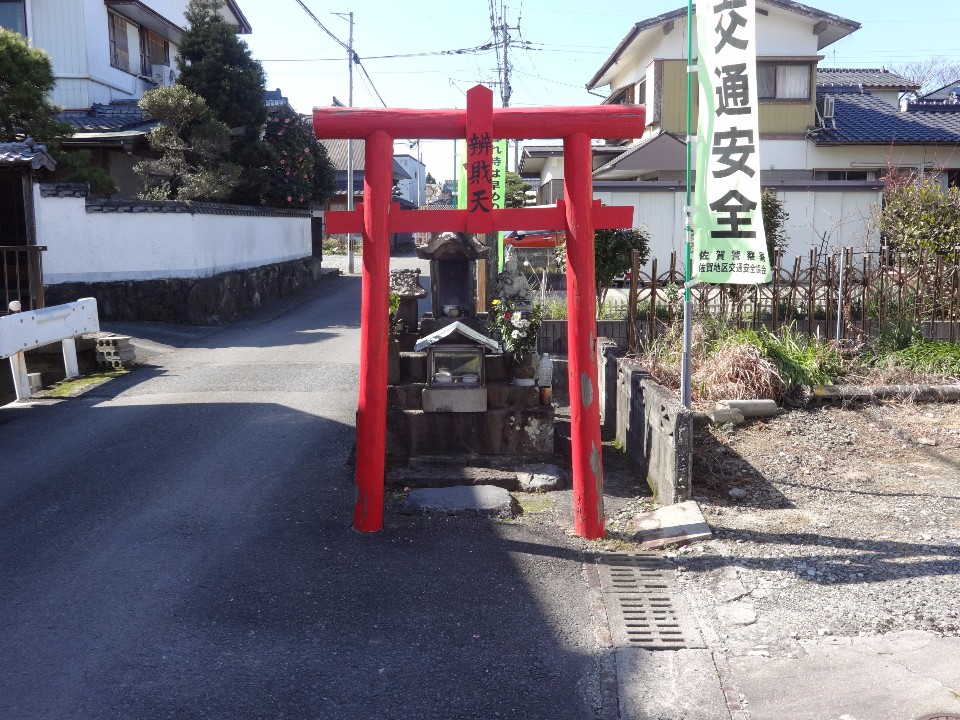 青空の下、住宅街の細い路地が分岐する場所に、「辨財天」と記された鮮やかな赤い鳥居が佇み、その奥に小さなお堂や石碑が祀られている風景写真