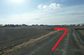 晴れ渡る空の下、広大な農地の中を貫く一本の未舗装路において、前方の交差点を左へと曲がる進路を強調するように、路面に沿って大きな赤い矢印が合成されている、ルート案内を目的とした風景写真