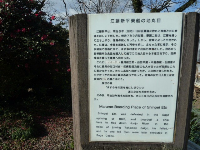 冬の柔らかな光が差し込む庭園で、赤い花を咲かせた椿の生垣を背景に、「江藤新平乗船の地丸目」という表題でその歴史的経緯と辞世の句、さらに英語での解説が併記された白い説明看板を正面から捉えた写真