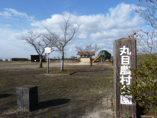 青空の下、手前に「丸目農村公園」と記された木目調の大きな看板が立ち、奥には滑り台などの遊具や小さな東屋が点在する、広々とした公園ののどかな風景を捉えた写真