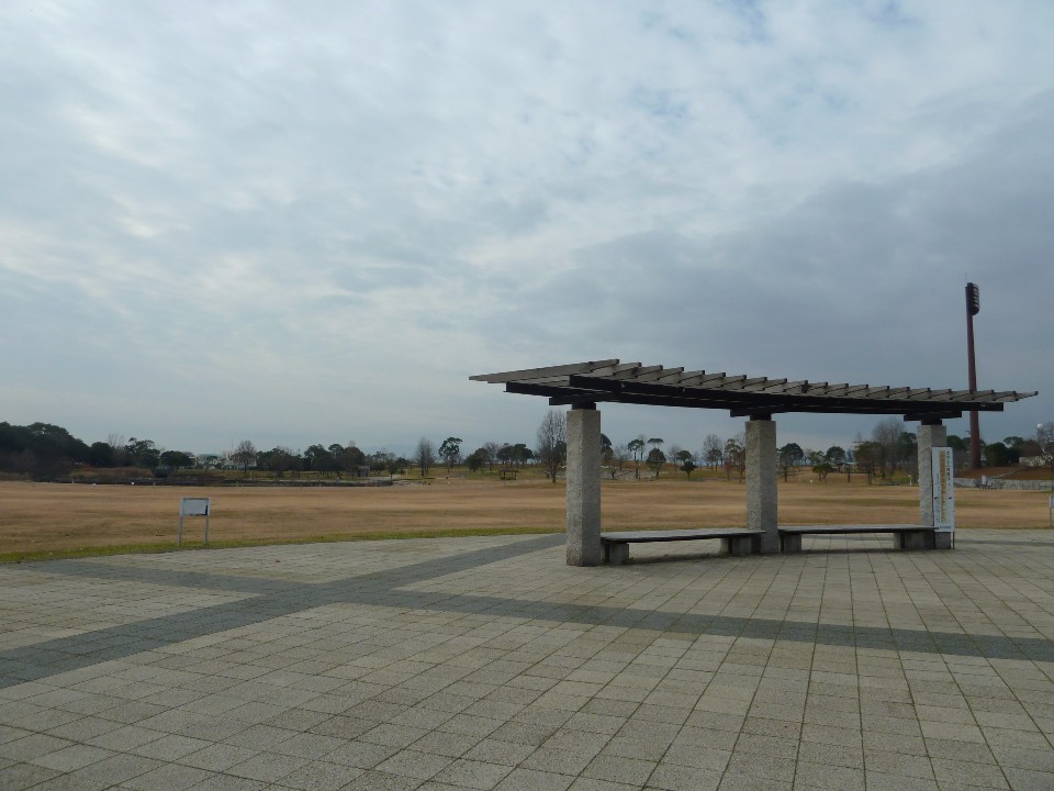 曇り空の下、広々とした芝生広場の手前に石造りの柱と平らな屋根を持つ東屋が設置され、地面は幾何学模様の石畳が広がる静かな公園の風景を捉えた、奥行きのある構図が特徴的な風景写真