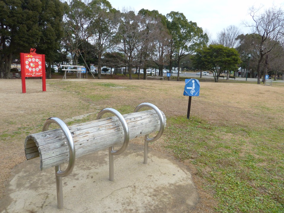 冬の枯れ草が広がる公園の広場に、木製の丸太を金属製の輪で固定した健康遊具が設置され、背後には赤い案内板や青いピクトグラムの看板、そして葉を落とした木々が並ぶ静かな冒険遊びの広場の写真