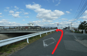 青空の下でガードレール沿いの坂道を上り、その先にある橋を渡って左方向へ進む経路を強調するように、路面上に大きく太い赤色の矢印が書き込まれた、周辺の民家や電柱が映り込む屋外の道路風景の写真