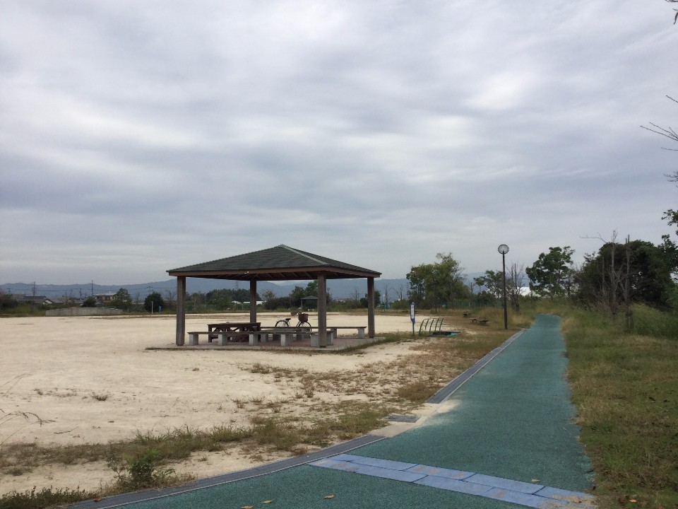 曇り空の下、広い砂地の広場に設置された東屋と木製のベンチ、その傍らに停められた自転車や奥へと続く青緑色の遊歩道、遠くに見える山並みや周囲の木々が静かな公園の風景を映し出している写真