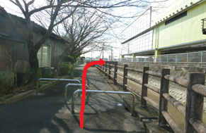 左側に木々や建物、右側に木製の柵と水路がある遊歩道において、路面に設置された金属製の車止めを越えて直進し、その先を右へと曲がる進行方向を赤い矢印で強調して示した写真