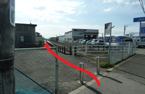 左側にフェンス、右側に並ぶ2本の金属製ポールと店舗の駐車場がある舗装路において、ポールの間を通り抜けて奥へと続く細い道へ直進する進行方向を、路面上に太い赤色の矢印を重ねて強調した写真